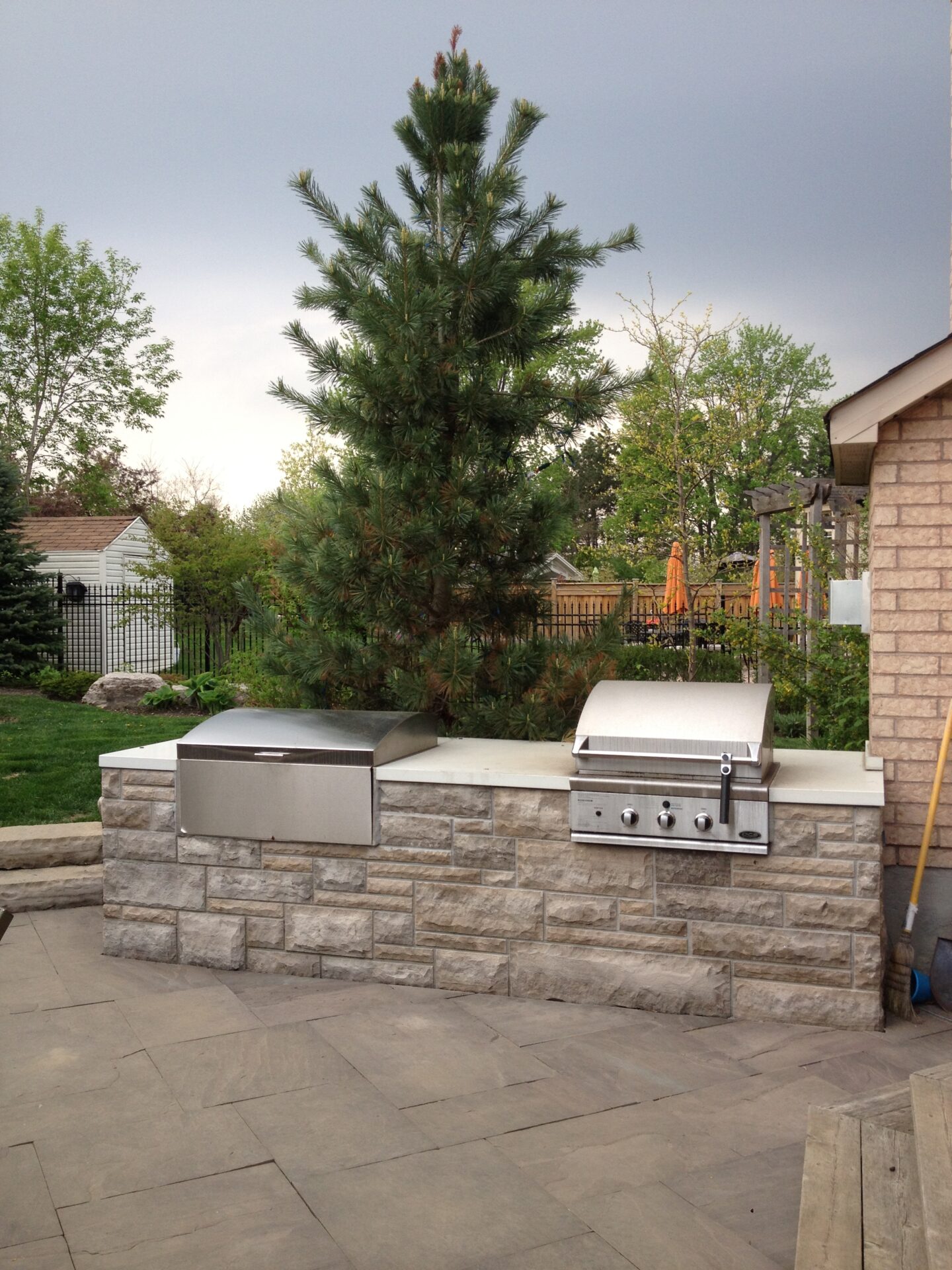 Outdoor stone grilling area with a built-in barbecue sits under a large pine tree. Surrounding greenery and wooden structures in the background.