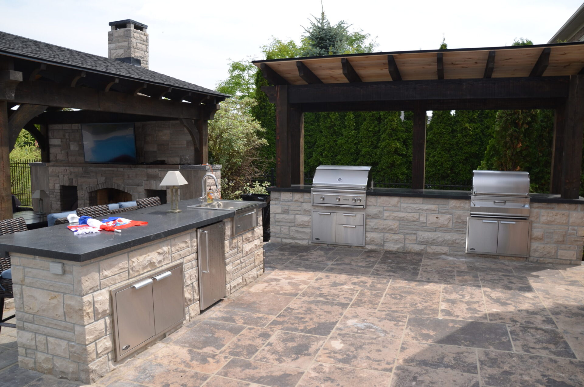 Outdoor kitchen with stone countertops, stainless steel appliances, and wooden pergola. Features a built-in grill and seating area in a backyard.