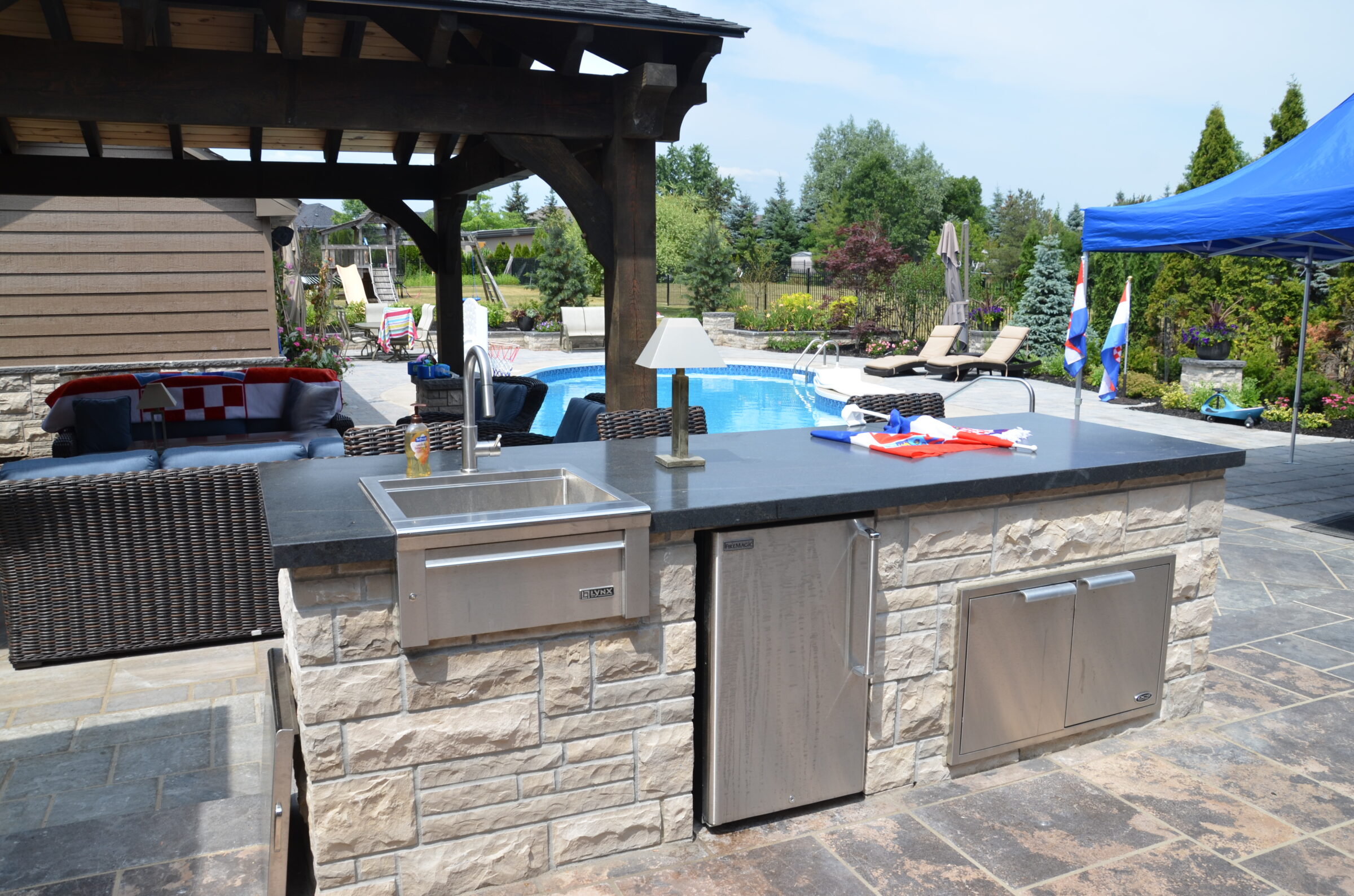 Outdoor kitchen with stone counters, sink, and grill near a pool. Patio furniture under a gazebo, surrounded by lush greenery and blue sky.