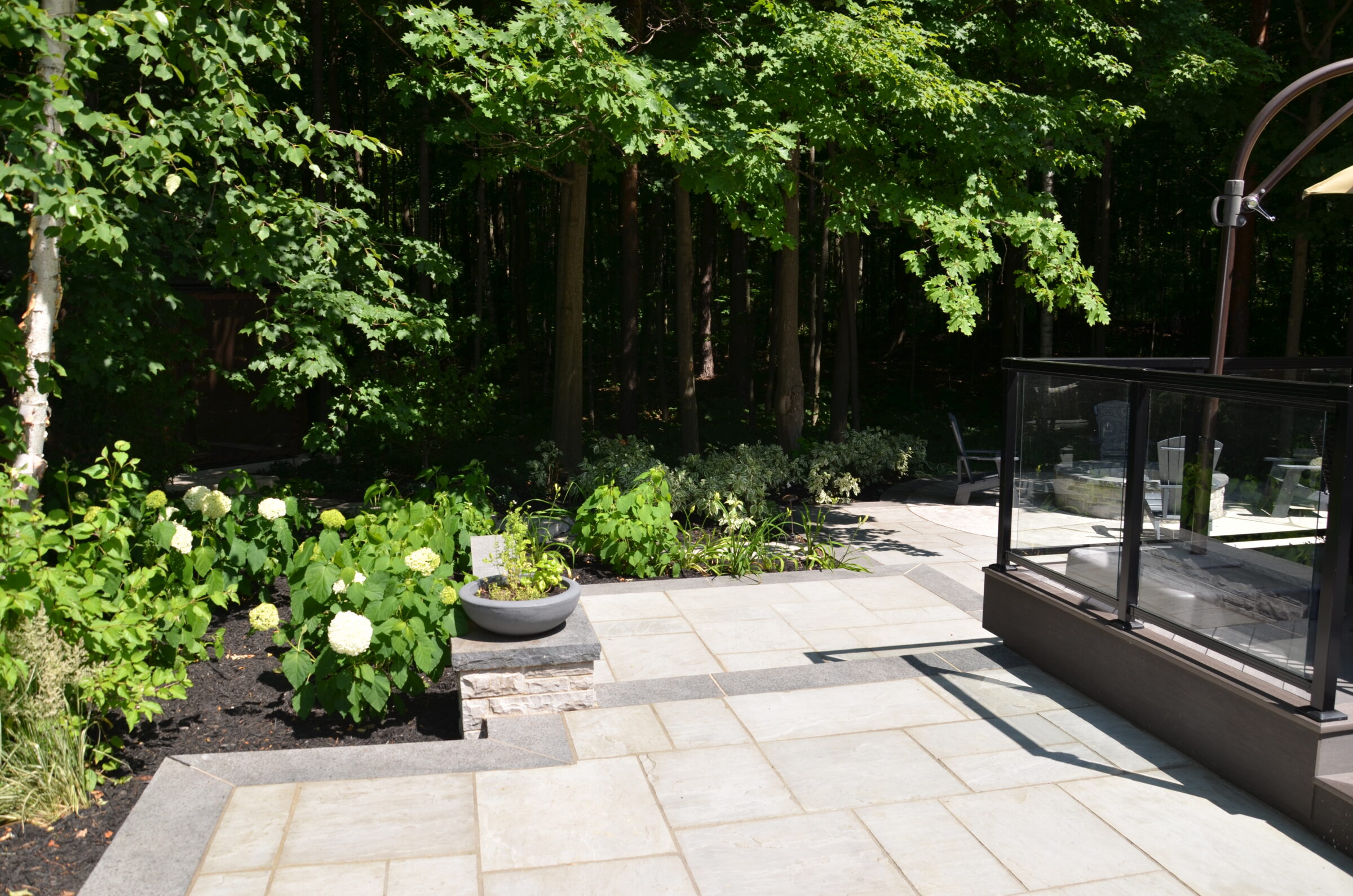 A sunlit patio with stone tiles, surrounded by lush greenery and potted plants, adjacent to a glass railing and wooded area.