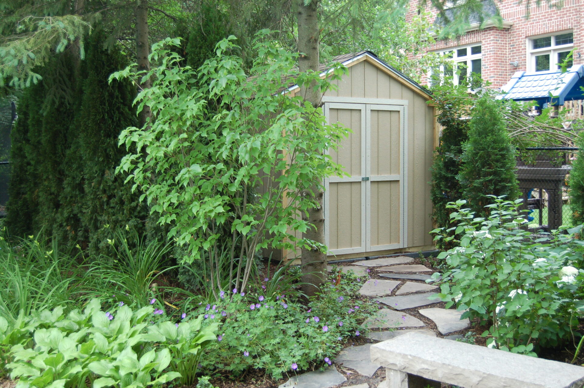 A small garden with lush greenery surrounds a wooden shed. Stone path leads to it, with a brick building partially visible in the background.