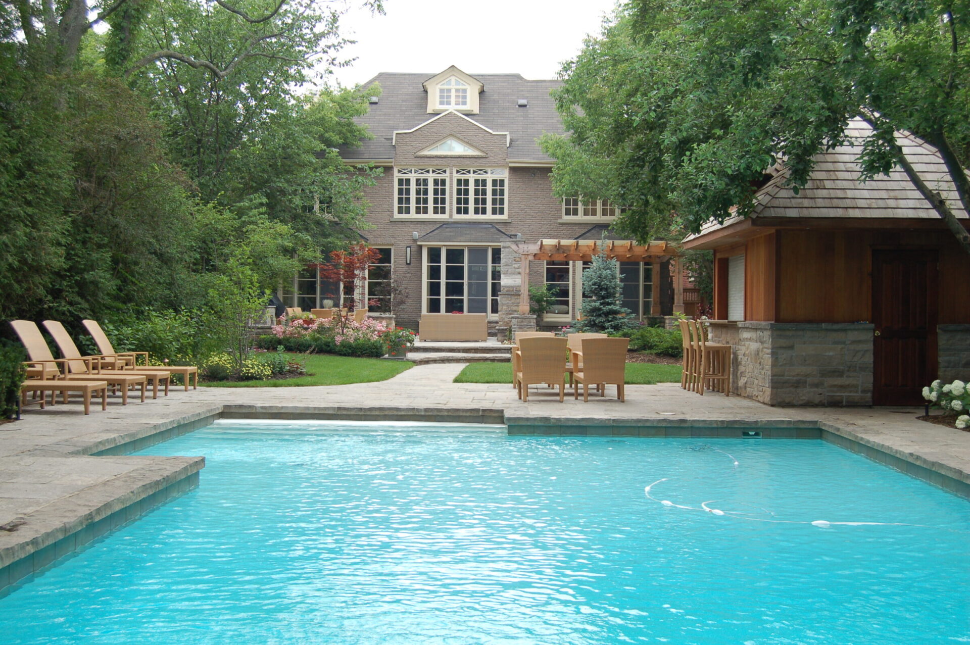 A serene backyard features a sparkling swimming pool, surrounded by lush greenery, a wooden gazebo, and a large, elegant brick house with tall windows.