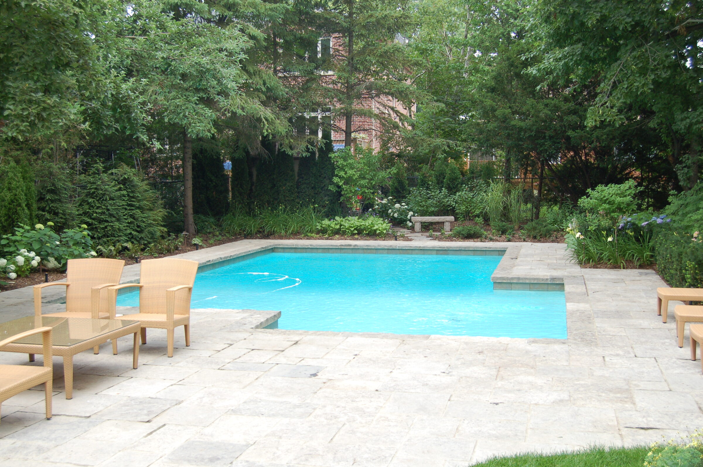 A serene backyard features a clear blue swimming pool, surrounded by lush greenery and outdoor furniture on a stone patio. No people visible.