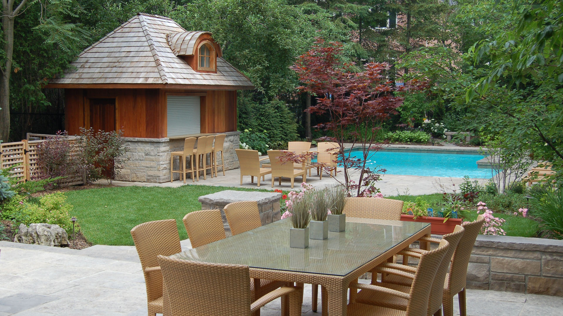 A serene backyard features a wooden pool house, lush greenery, a swimming pool, and a patio with a glass table and wicker chairs.
