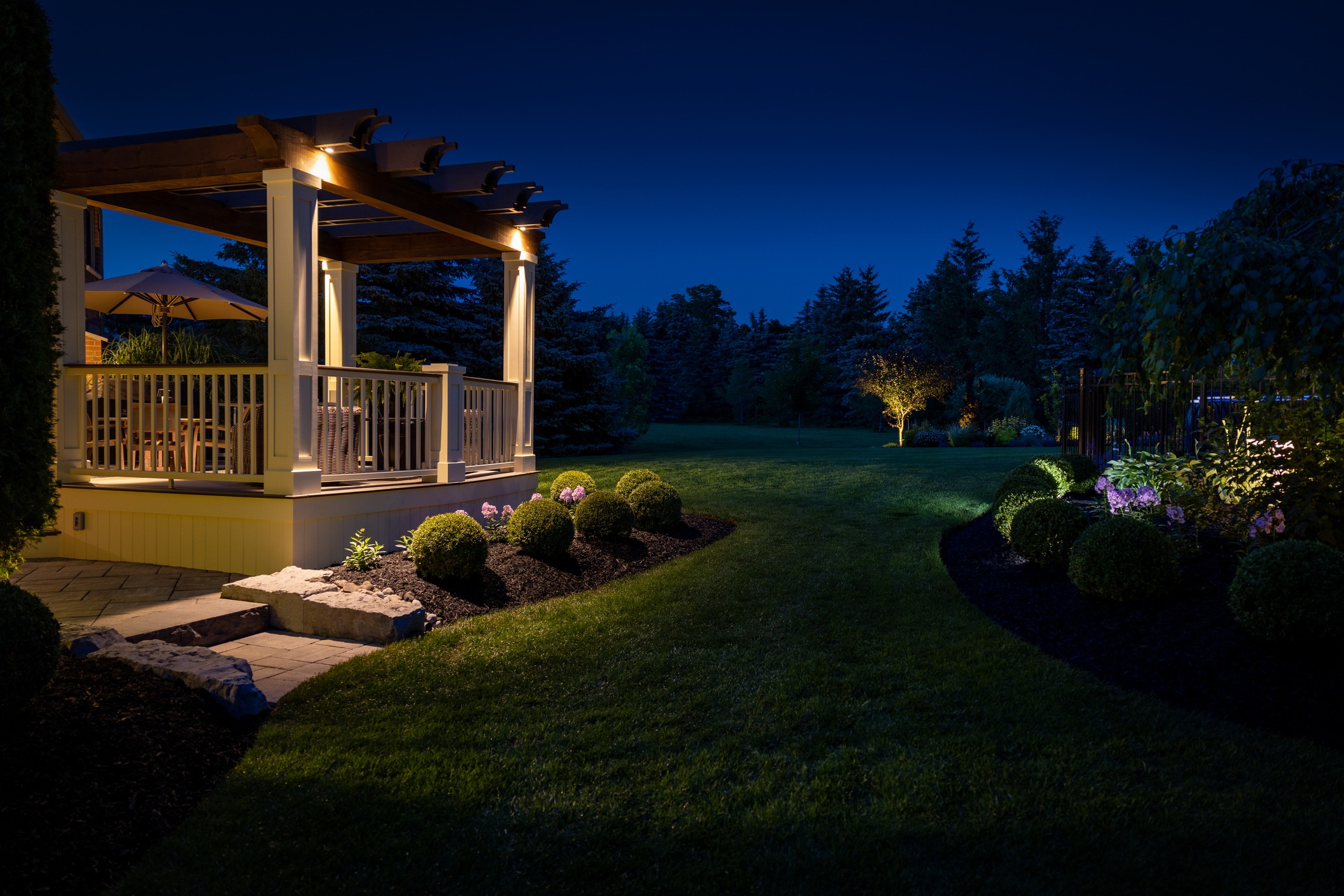 A well-lit backyard at night features a wooden pergola and patio, with manicured landscaping and garden lights illuminating trees and shrubs.