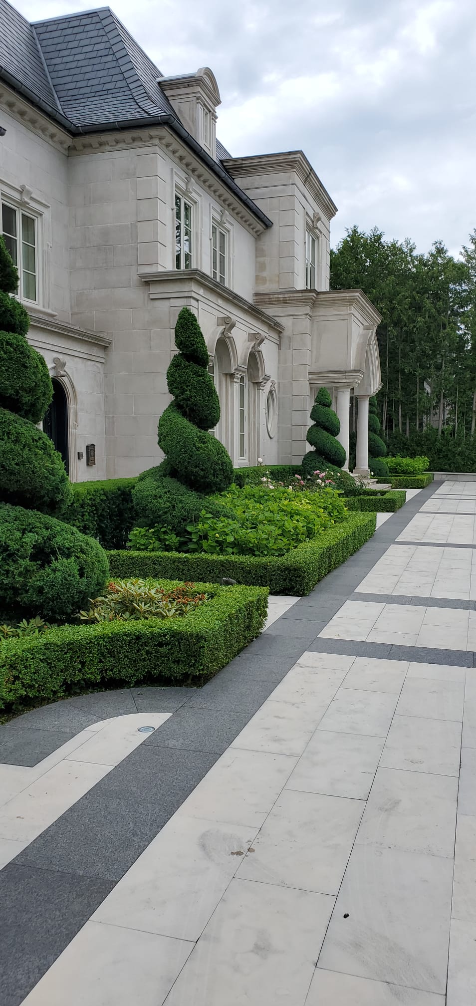 A grand stone mansion with manicured hedges and spiral topiaries lines a checkered pathway, set against a backdrop of lush trees.