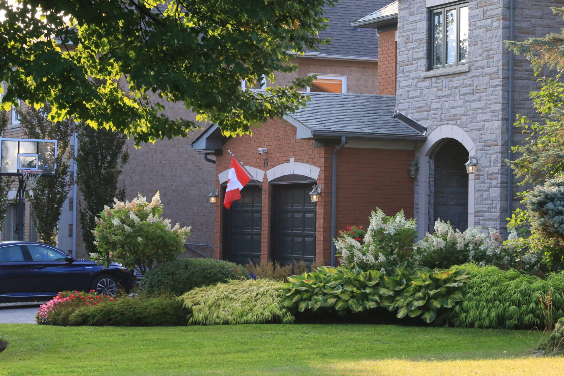 A suburban home with a Canadian flag, surrounded by lush greenery and a parked car. The house features brick and stone details.