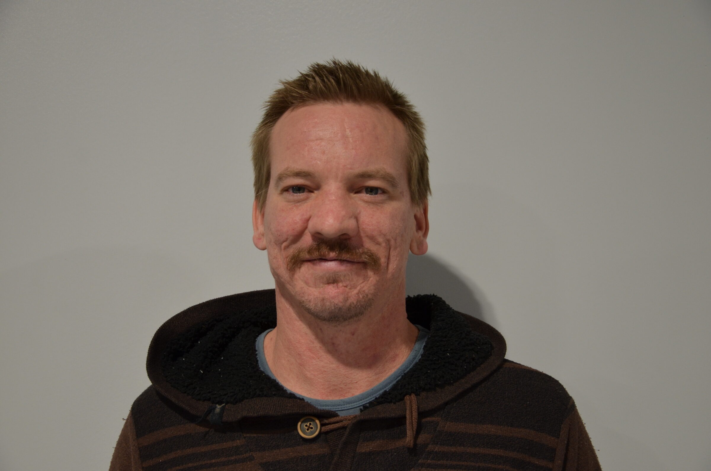 A person wearing a dark hoodie smiles slightly against a plain, light gray background. The lighting is soft, casting minimal shadows.