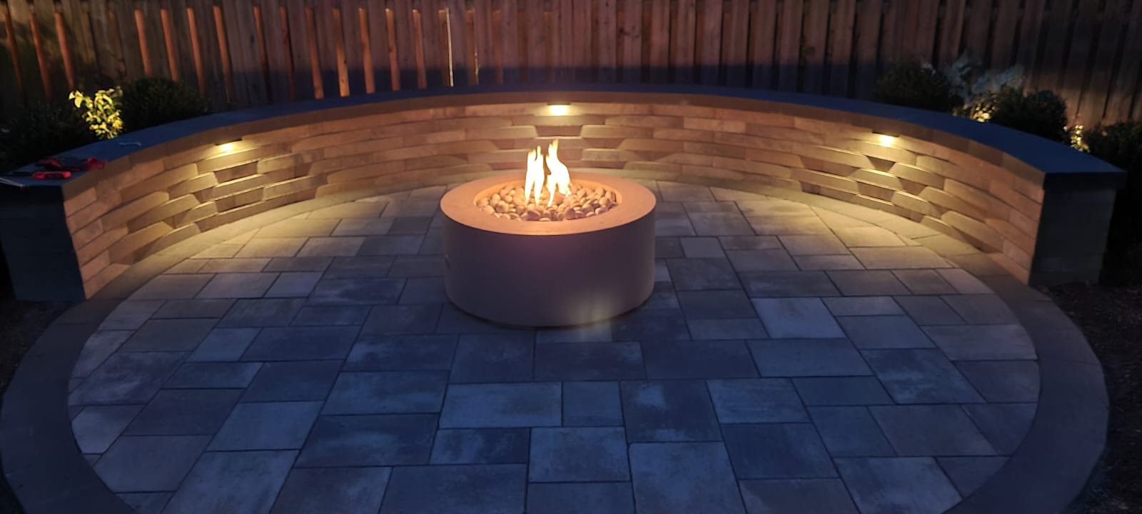 A circular patio with a central fire pit, surrounded by a stone bench and illuminated by small wall lights, creates a cozy ambiance.