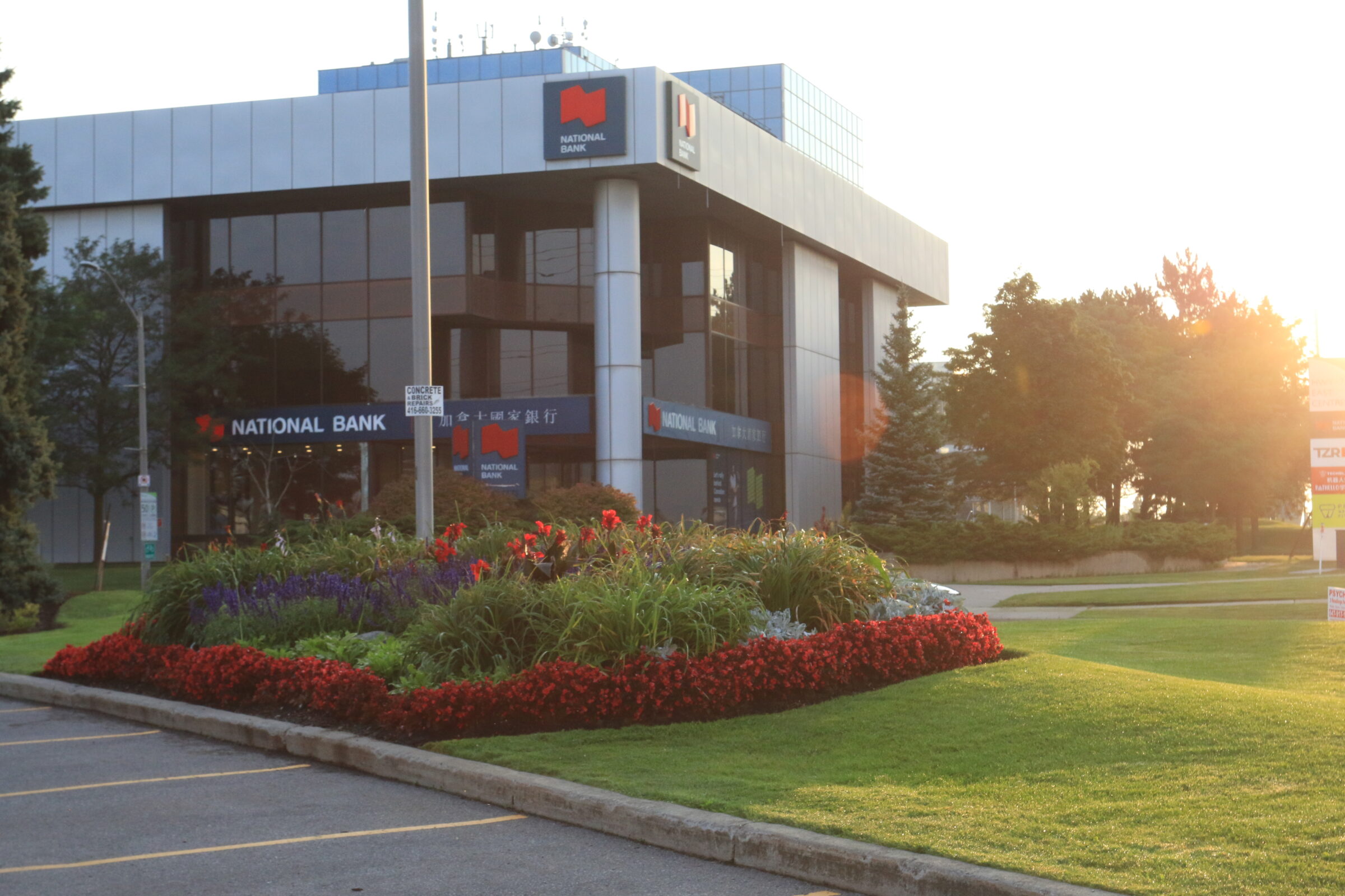 Modern National Bank building with reflective glass, surrounded by vibrant flowerbeds and neatly manicured lawns, bathed in warm, late afternoon sunlight.