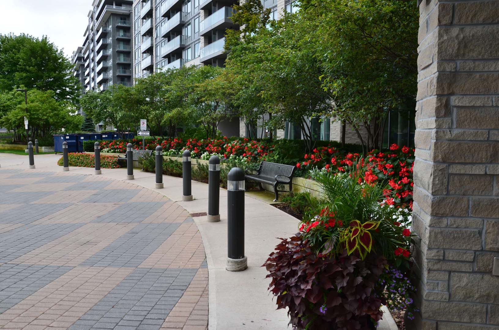 A modern apartment complex is surrounded by lush greenery, vibrant red flowers, and a pathway with stone columns and bollards, creating a serene urban setting.