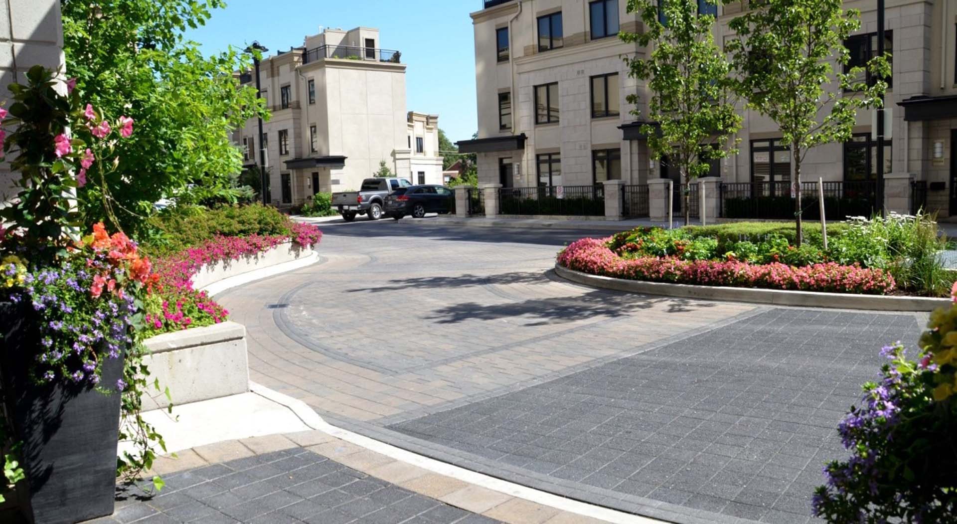 A landscaped courtyard with elegant buildings, vibrant flowers, and a paved driveway. Trees line the area, enhancing the tranquil, urban setting.