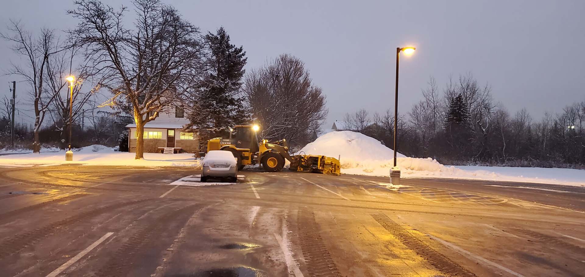 A snowy parking lot features a snowplow clearing snow. A lit building and bare trees are visible under the dusky sky.
