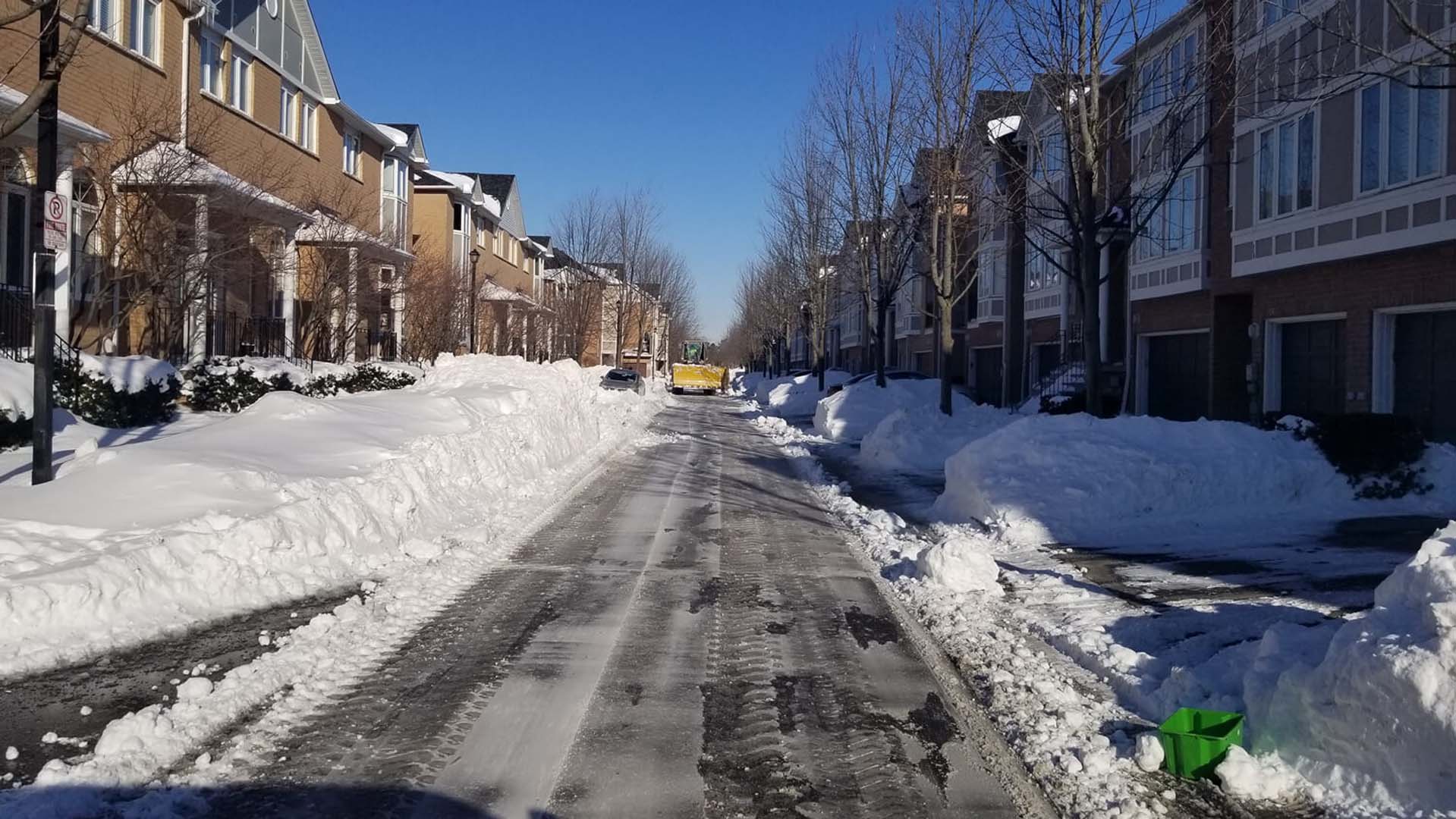 Snow-covered residential street with cleared paths, flanked by modern townhouses. Bright blue sky and bare trees line the road. No visible landmarks.
