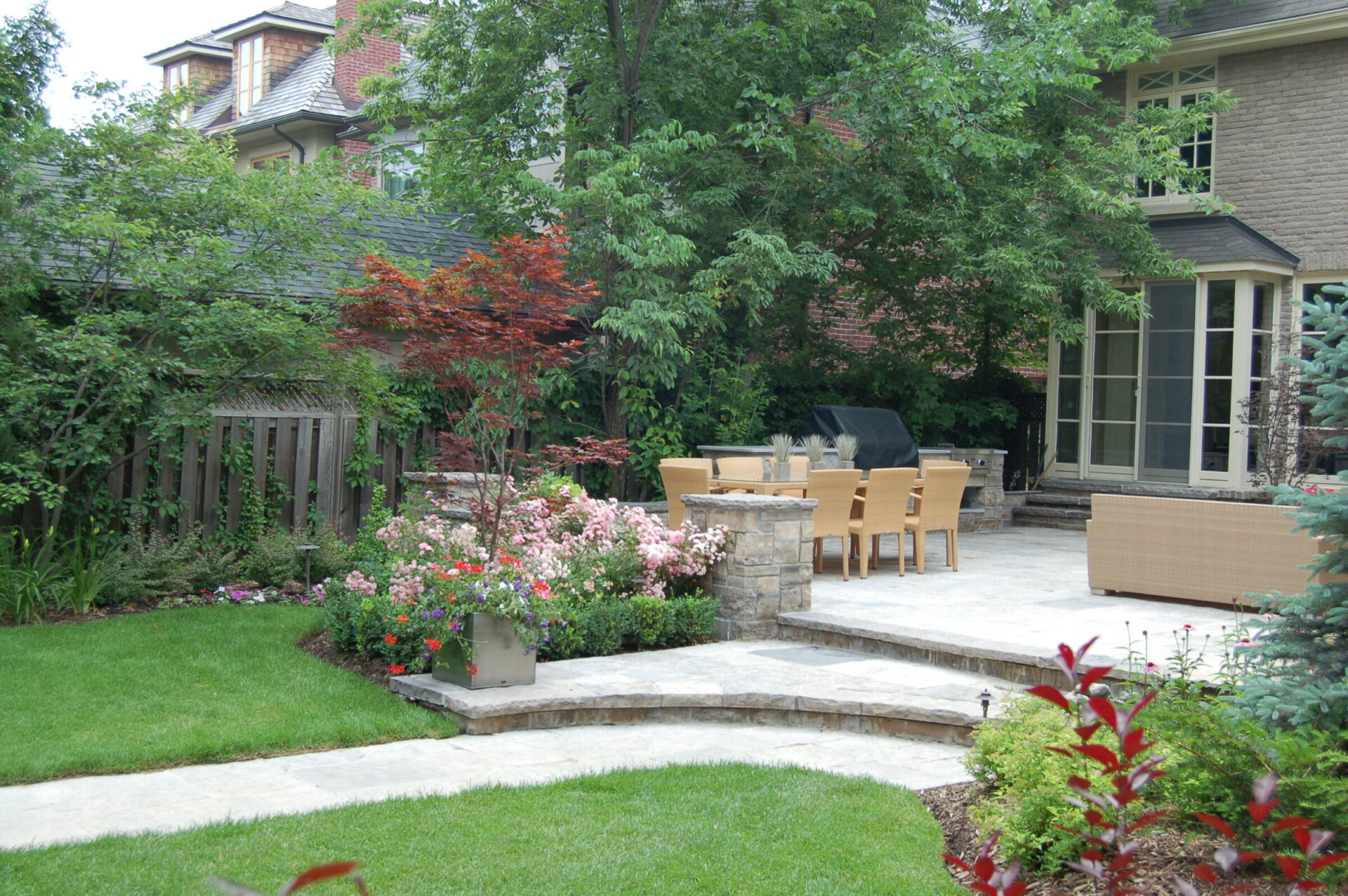 A well-maintained garden with a patio features dining furniture, lush greenery, and colorful flowers. A stone pathway leads through the vibrant outdoor space.