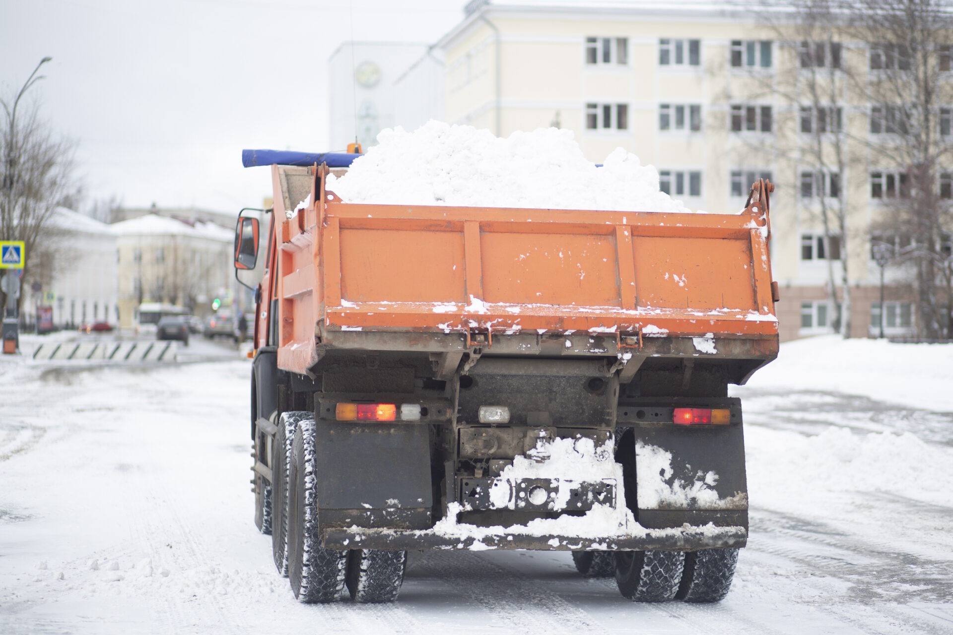 An orange truck loaded with snow is on a snowy street, with a multi-story building and bare trees in the background.