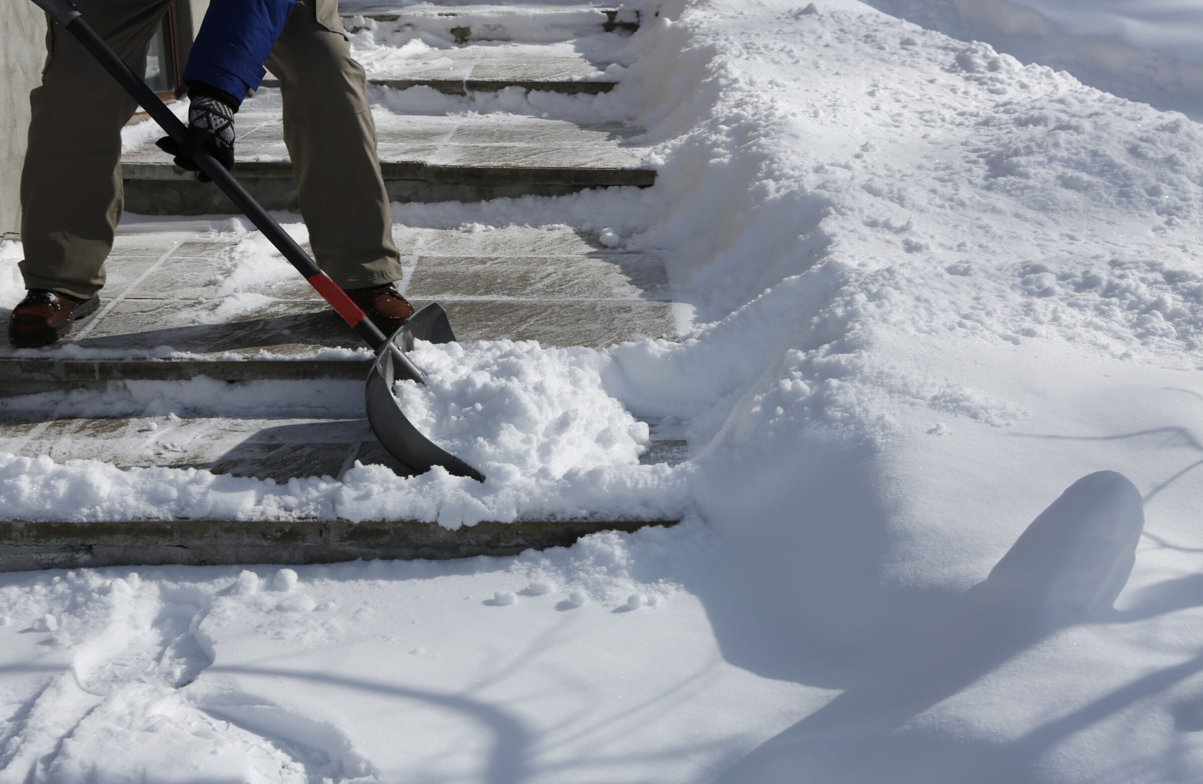 A person shoveling snow from outdoor steps, wearing winter clothing. Snow is piled on both sides, suggesting recent snowfall. Shadow cast by a shovel.