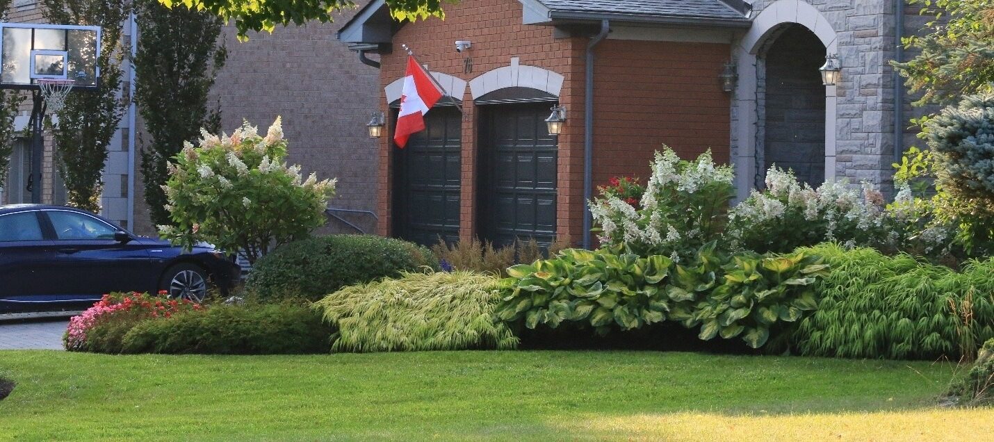 A suburban house with a Canadian flag, surrounded by lush greenery and trees. A parked car is visible in the driveway.