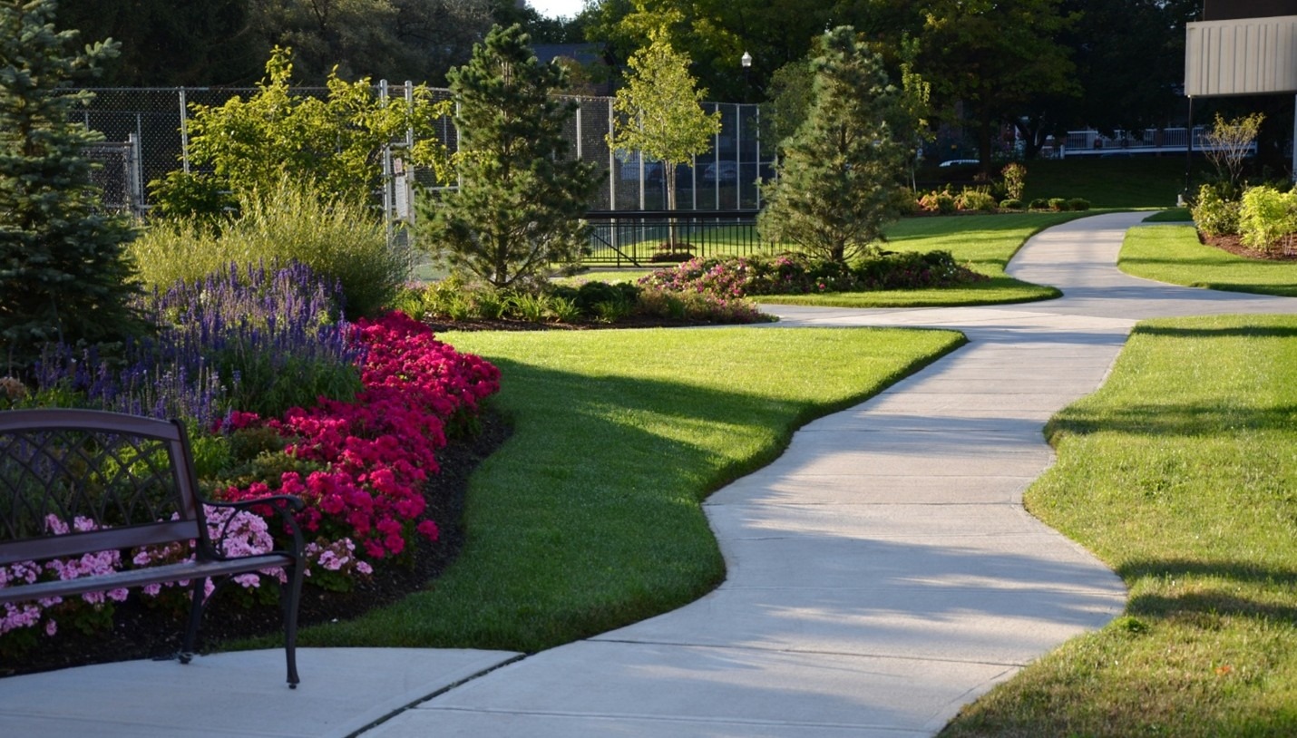 A winding path through a well-maintained garden with colorful flowers and trees, adjacent to a bench and an enclosed tennis court.