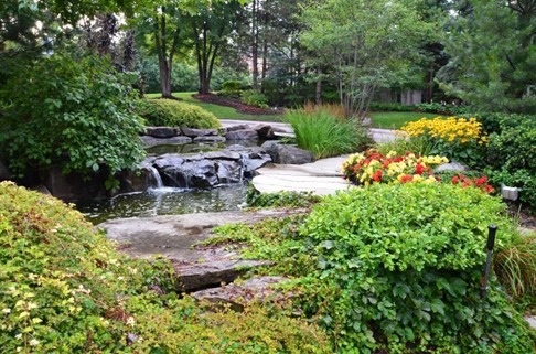 A tranquil garden features a small waterfall, surrounded by lush greenery, vibrant flowers, and a stone pathway, creating a peaceful natural setting.