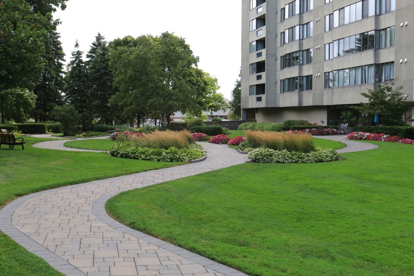 A winding stone path weaves through a well-maintained garden beside a modern apartment building, featuring lush trees and vibrant flower beds.