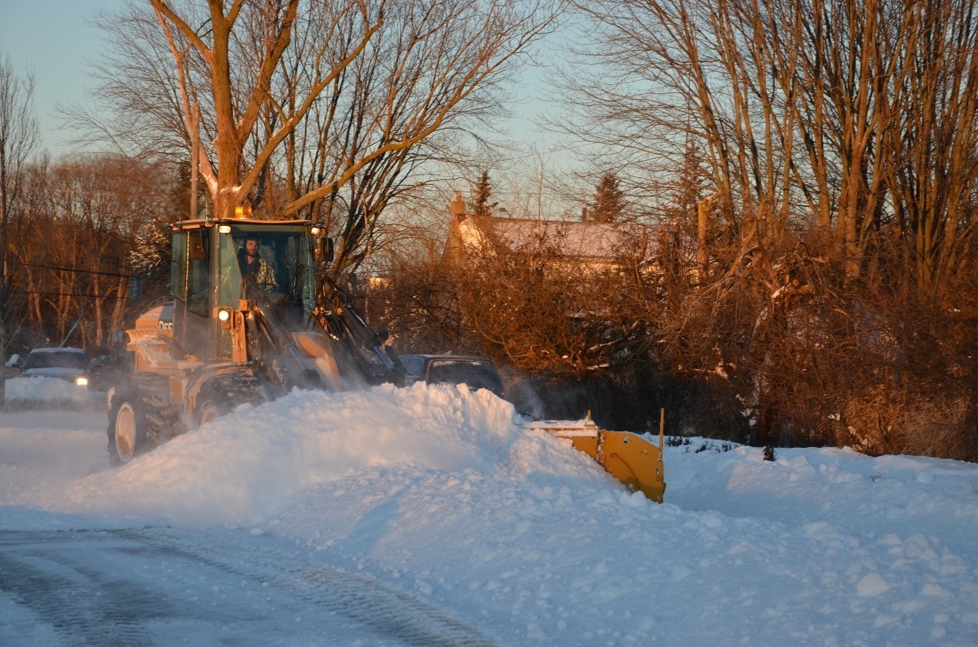 A snow plow clears a street in a snowy residential area, surrounded by leafless trees and houses, with a car following behind.