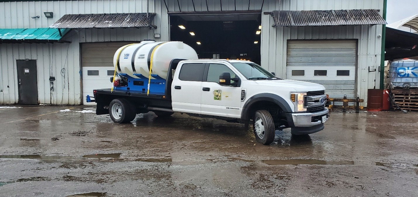 A white utility truck carrying large tanks is parked in front of an industrial garage on a wet, overcast day.