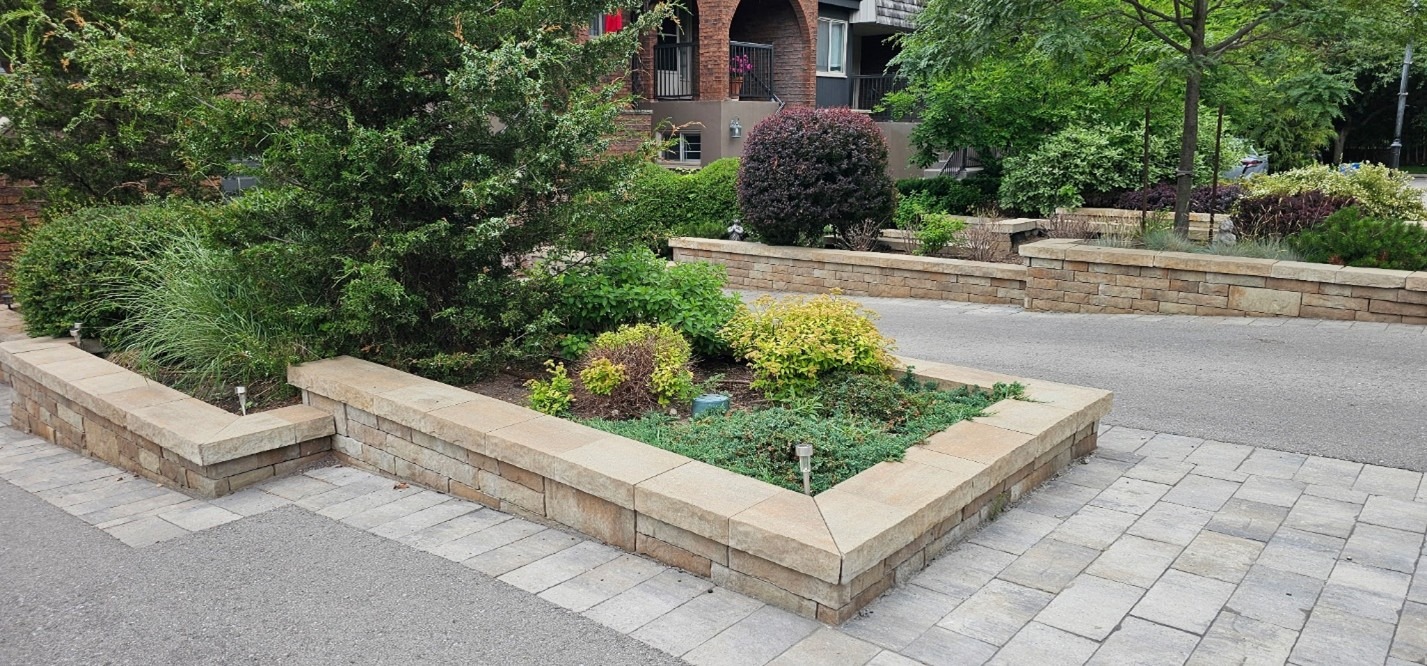 A landscaped garden with various shrubs and trees, bordered by stone walls, alongside a paved pathway and a brick building in the background.