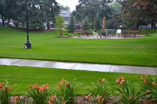 A well-maintained park with lush grass, flower beds, and seating area under an umbrella. Overcast sky and pathway leading through the greenery.