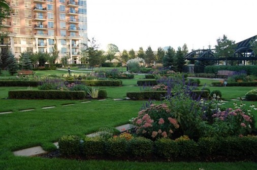 A lush garden with colorful flowers and trimmed hedges, adjacent to a modern building. Several people stroll on a pathway.