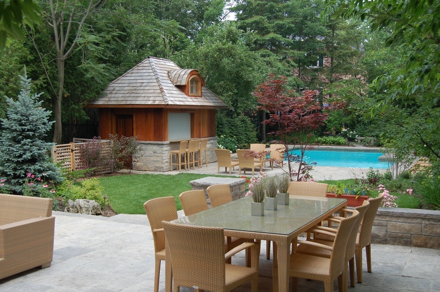 A backyard with a patio features a dining table, chairs, a small wooden house, lush greenery, and a pool surrounded by trees.