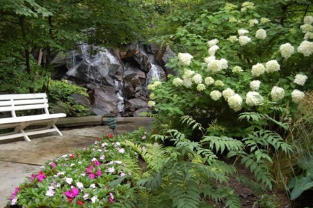 A serene garden features lush greenery, blooming flowers, a white bench, and a small waterfall nestled among rocks and trees.