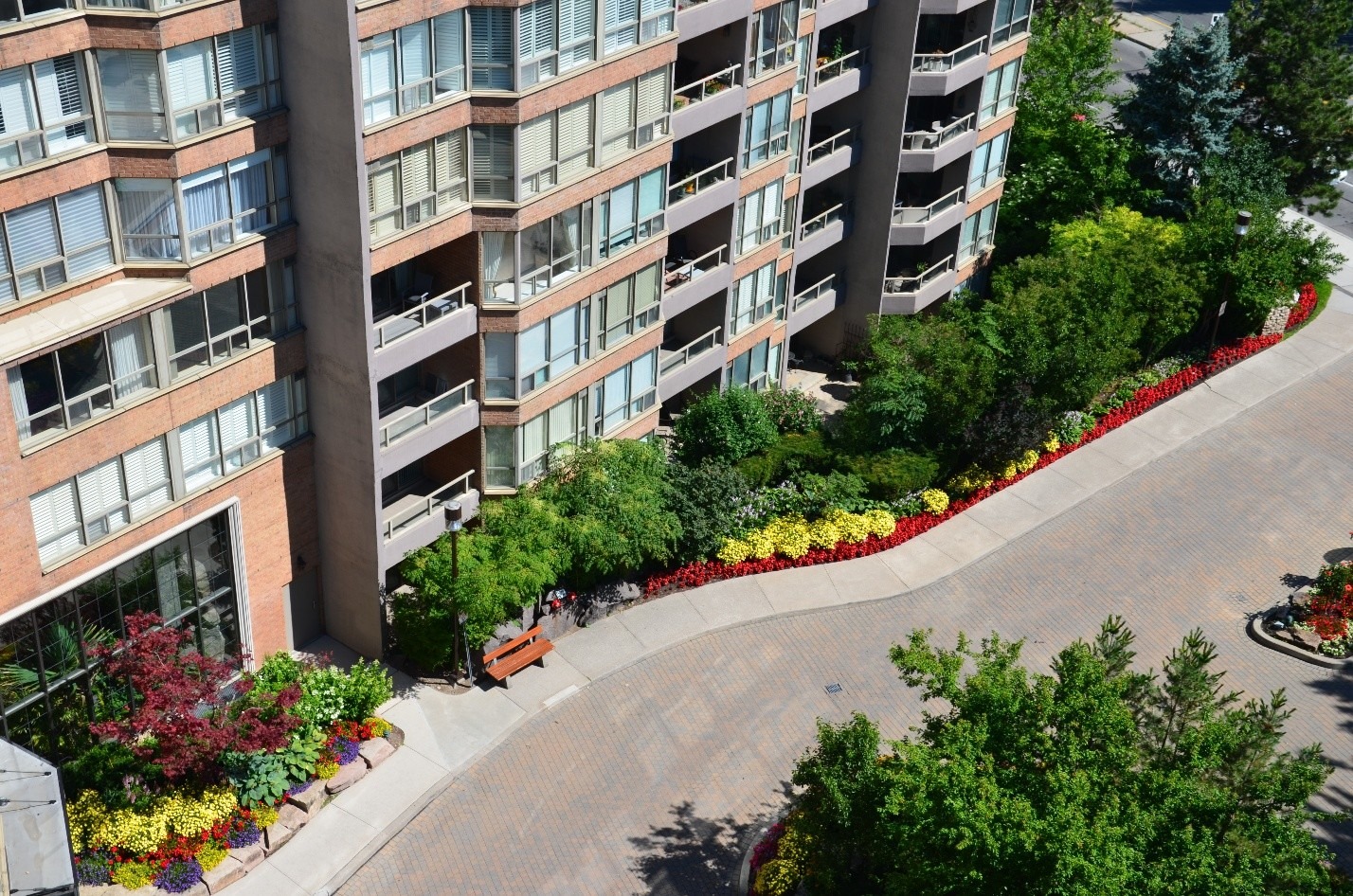 An apartment building with lush gardens and colorful flowers lining a curved walkway. A person sits on a bench near the building.