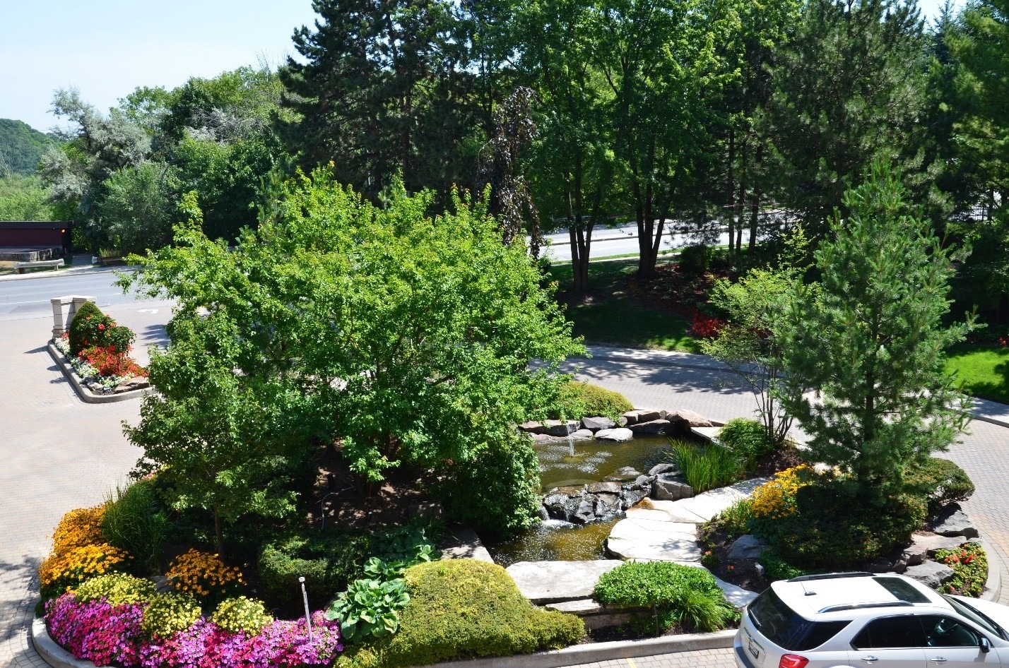 A landscaped garden with colorful flowers surrounds a small pond, near a parked car, bordered by trees and a road in the background.