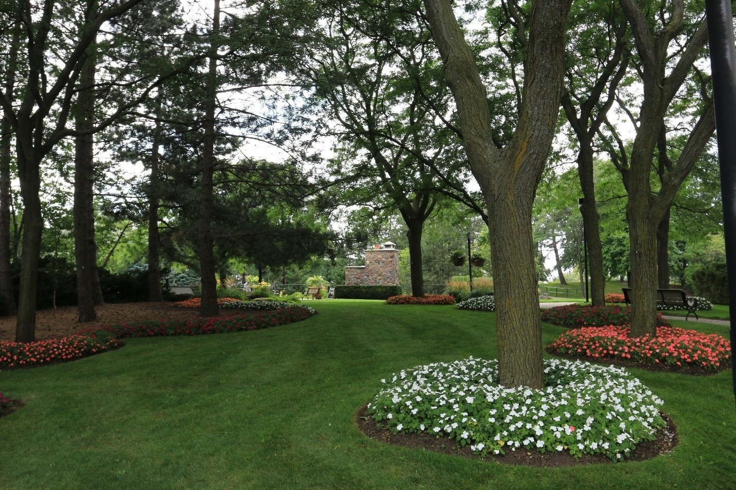 A landscaped garden with neatly mowed grass, vibrant flower beds, and tall trees. A brick structure is visible, surrounded by lush greenery.
