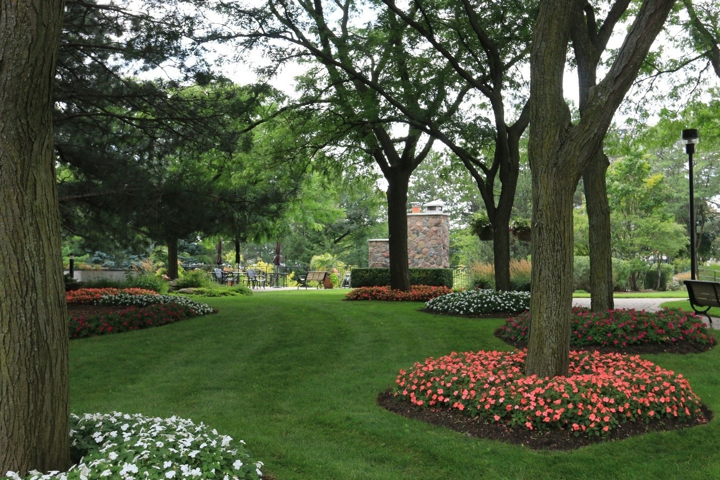 A lush, green park scene with blooming flower beds, tall trees, and a stone structure. No visible people or landmarks present.