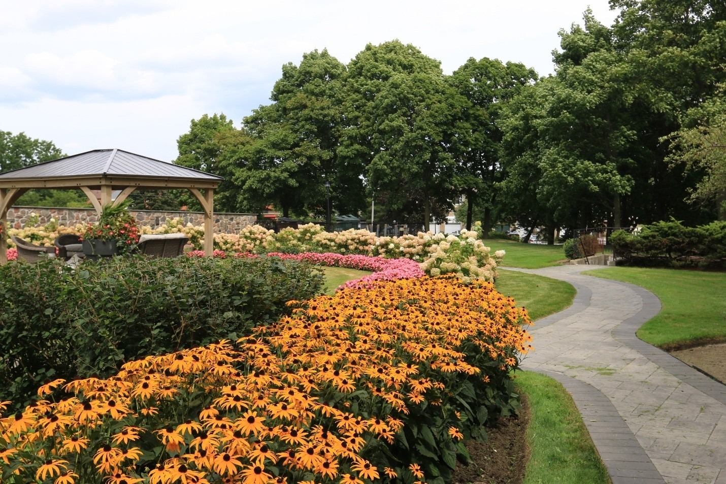 A garden with vibrant orange flowers and a gazebo, surrounded by lush greenery and a winding path, under a partly cloudy sky.