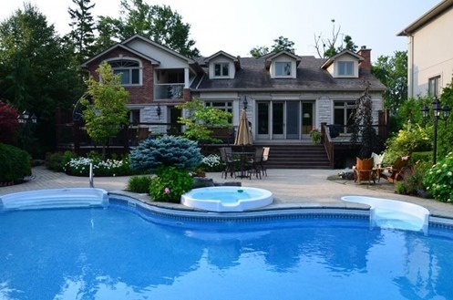 A spacious backyard features a luxurious pool, hot tub, and patio in front of a large house surrounded by lush greenery.