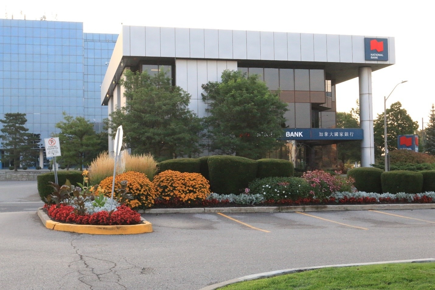Modern bank building with reflective glass, surrounded by manicured gardens and vibrant flowers. Clear signage and a well-maintained parking lot in the foreground.