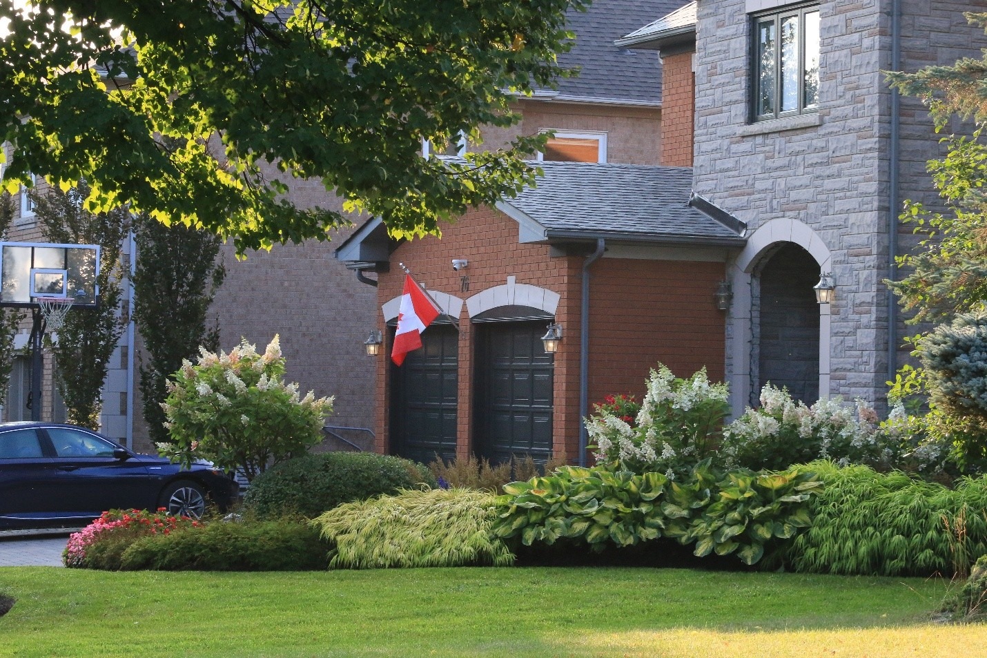 Suburban house with a Canadian flag in the front yard. Lush greenery and a parked car enhance the peaceful, residential atmosphere.