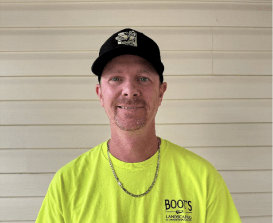 A person wearing a yellow "Boot's Landscaping" shirt and a black cap smiles against a beige paneled wall.