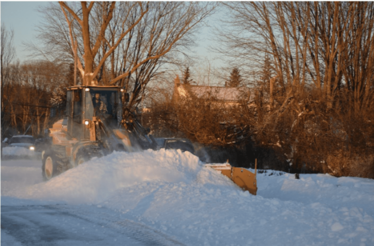 A snowplow clearing snow from a road surrounded by bare trees and a house in the background during a sunny winter day.