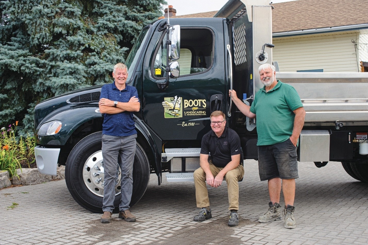 Three people pose by a landscaping truck labeled "Boots Landscaping," parked on a stone-paved driveway with trees and a house in the background.