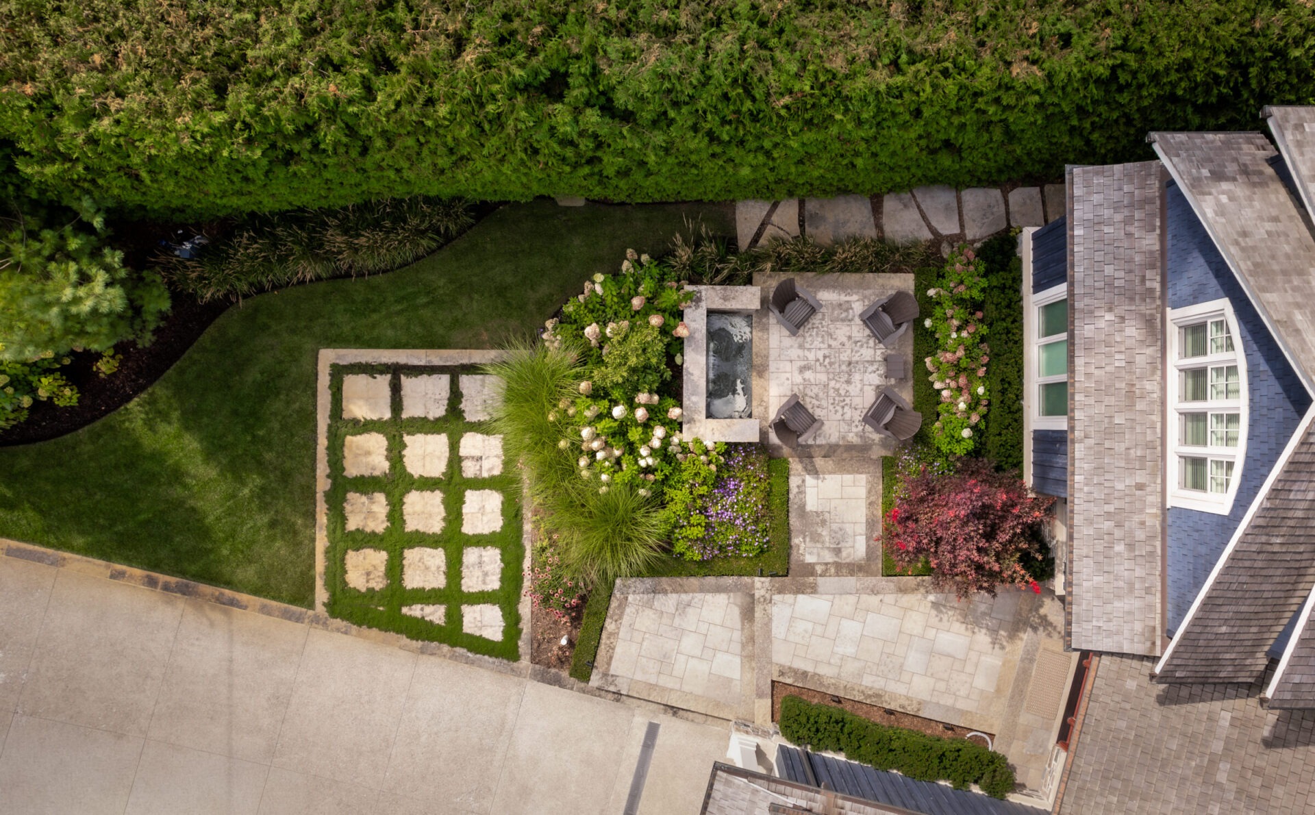Aerial view of a landscaped backyard featuring a patio, garden, and parking area, surrounded by lush greenery and a house.
