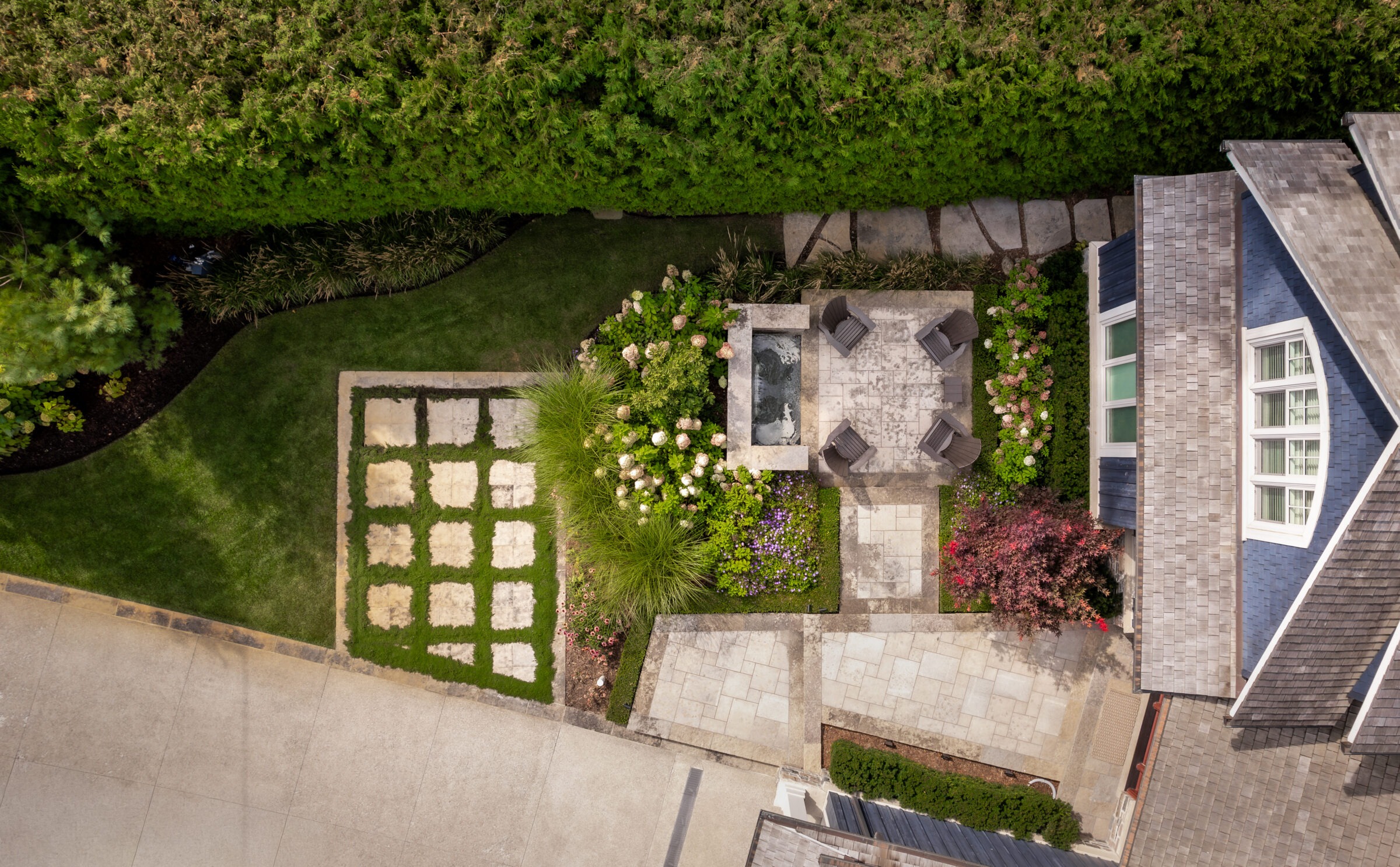 Aerial view of a landscaped backyard featuring a patio, garden, and parking area, surrounded by lush greenery and a house.