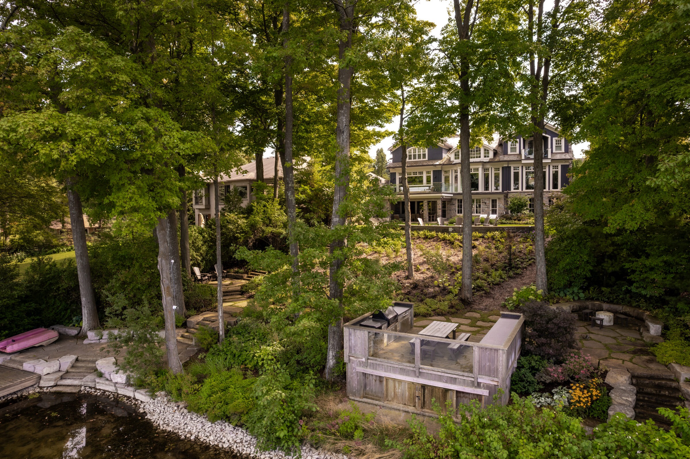 Lakeside scene featuring a modern house surrounded by lush trees, with a small dock and seating area by the water.