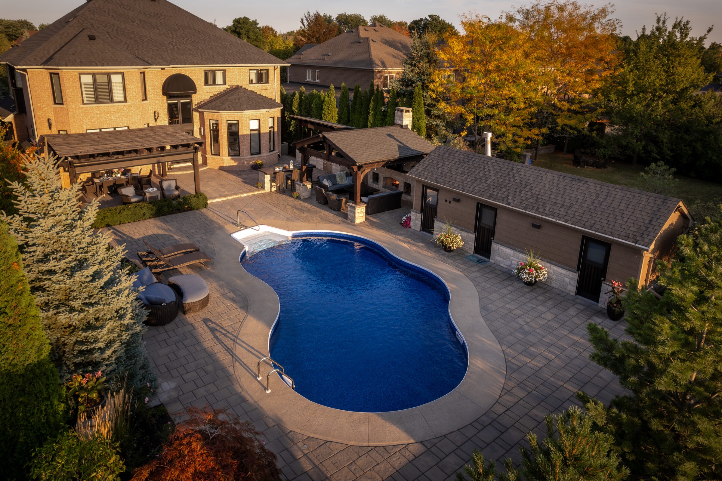Aerial view of a luxury house with swimming pool, patio, and garden surrounded by trees. Outdoor seating and decorative elements are visible.