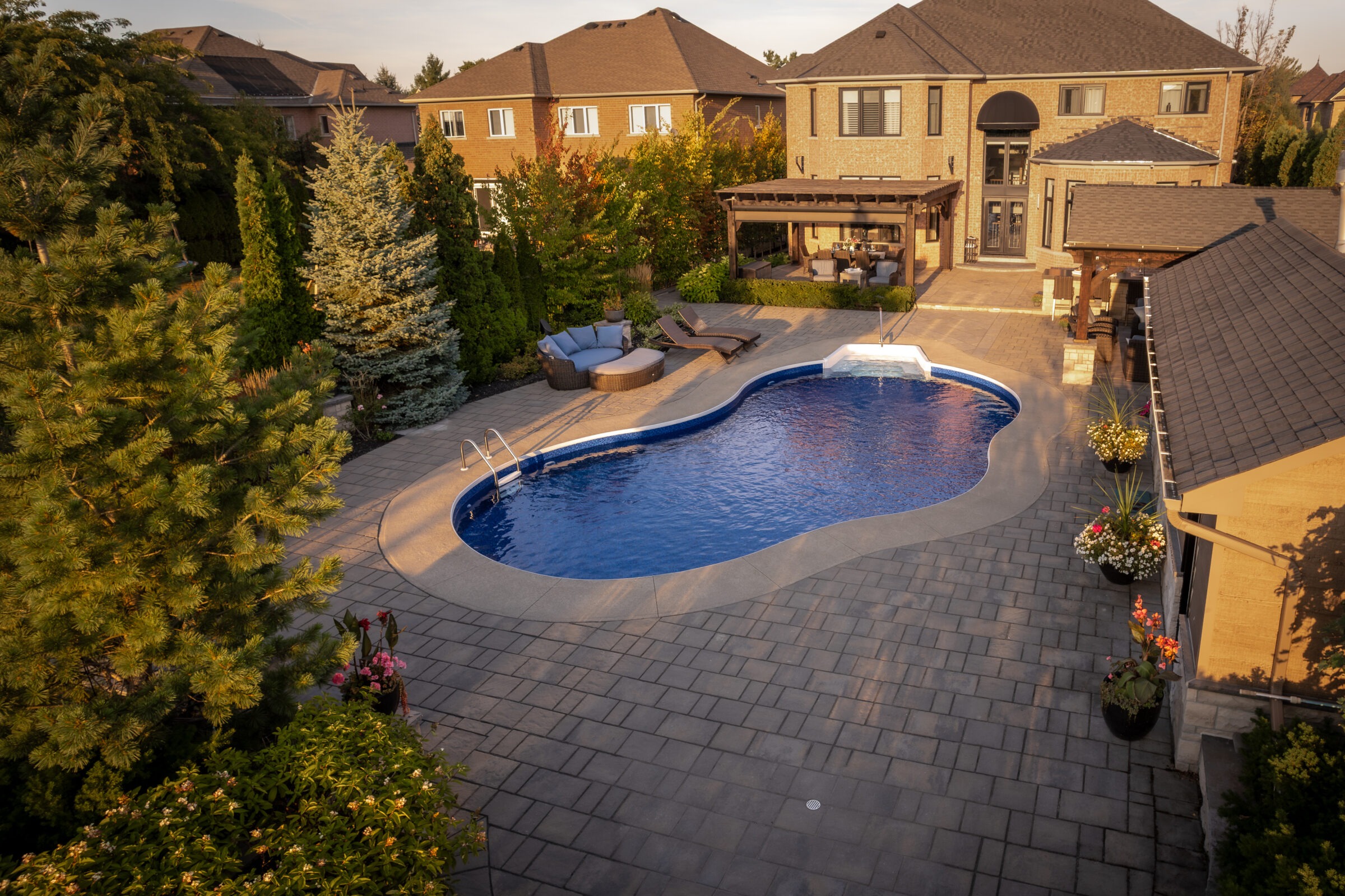 A backyard with a kidney-shaped pool, patio furniture, and landscaped garden, surrounded by houses. One person stands near the pool area.