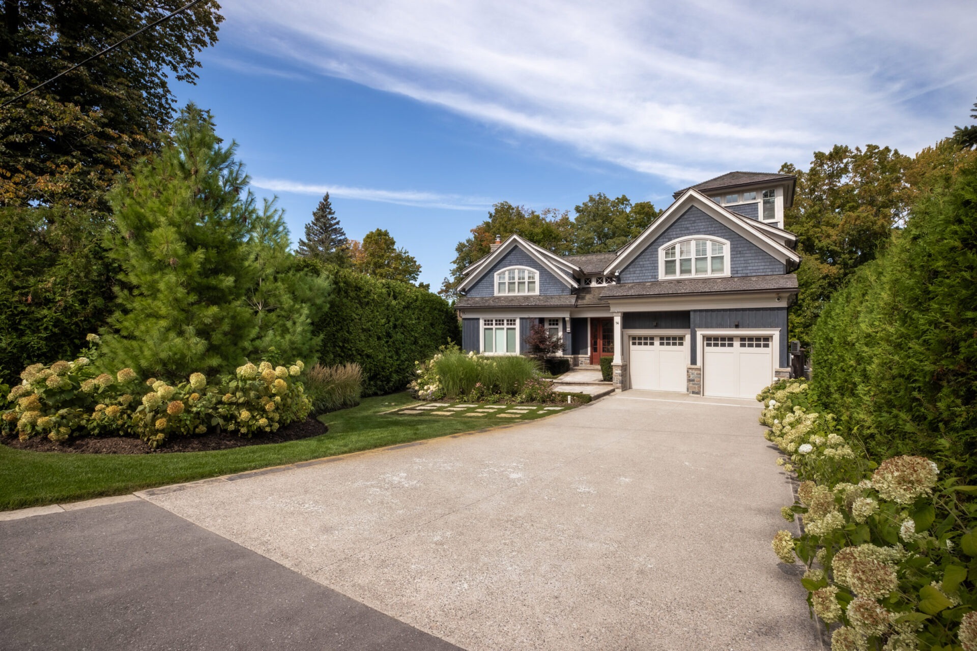 A suburban house with a two-car garage is surrounded by lush greenery. The front yard features landscaped plants and a paved driveway.