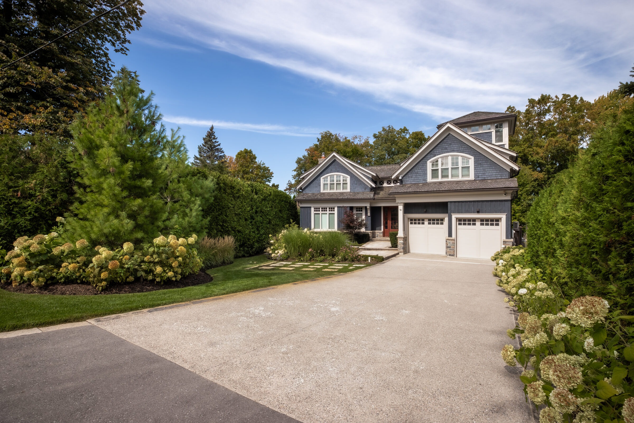 A suburban house with a two-car garage is surrounded by lush greenery. The front yard features landscaped plants and a paved driveway.